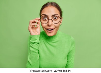 Photo of surprised cheerful woman with dark hair looks wondered through spectacles cannot believe her eyes dressed in casual jumper isolated over vivid green background. Human reactions concept