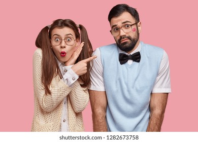 Photo of surprised European woman with two pony tails, red lipstick, points at hesitant boyfriend who recieved kiss from her, wears square glasses and elegant outfit, pose together in studio - Shutterstock ID 1282673758