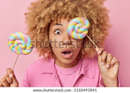 Photo of surprised young European woman with curly hair covers eye with muticolored lollipop keeps mouth opened from wonder dressed in jacket isolated over pink background. Sweet tooth concept