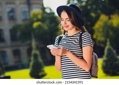 Photo of sweet cute young woman dressed striped outfit cap backpack writing message modern device smiling outside landscape