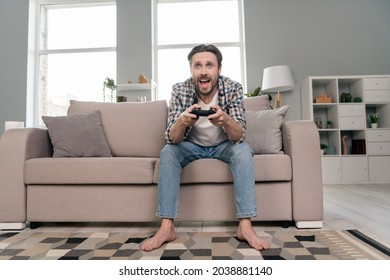 Photo of sweet funny young man wear plaid shirt sitting sofa holding playstation smiling indoors house room