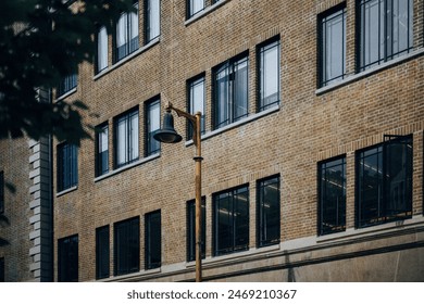 A photo of a tall brick building with a black metal fire escape and large windows. A street lamp with a curved glass shade is mounted on a black metal post in front of the building on the sidewalk. - Powered by Shutterstock