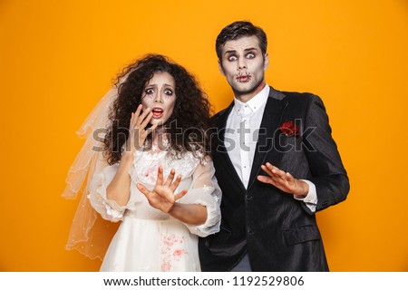 Photo of terrifying zombie couple bridegroom and bride wearing wedding outfit and halloween makeup scaring you isolated over yellow background