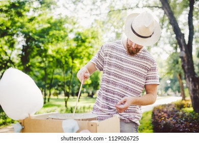 photo theme small business cooking sweets. A young man with a beard of a Caucasian trader in the hat the owner of the outlet makes candy floss, fairy floss or Cotton candy in the summer park.