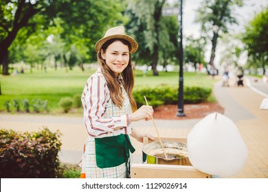photo theme small business cooking sweets. A young caucasian woman with an apron trader in the hat the owner of the outlet makes a candy floss, a fairy floss or a cotton candy in the summer park.