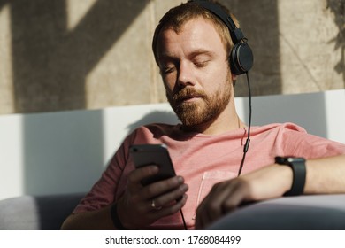 Photo Of Thinking Redhead Man With Beard Using Mobile Phone And Headphones While Sitting On Sofa At Home