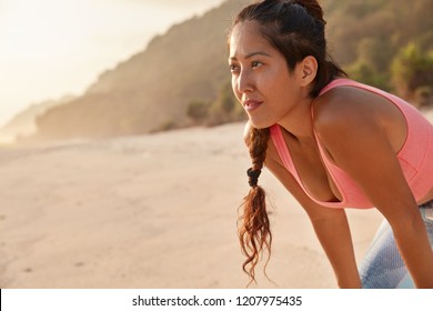 Photo of thoughtful Asian woman with pensive expression, leans at knees, focused into distance, has active cardio training, poses on beach, enjoys calm atmosphere with free space for slogan. - Powered by Shutterstock