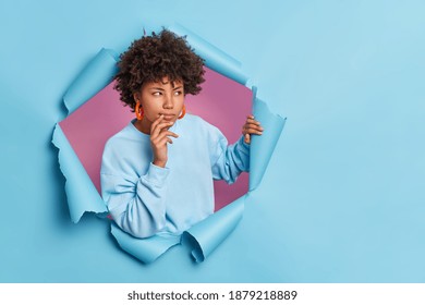Photo of thoughtful dark skinned Afro American woman looks away and considers something keeps hand near mouth breaks through paper hole thinks about decision wears casual jumper and earrings - Shutterstock ID 1879218889