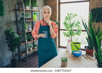 Photo of thoughtful doubtful senior woman dressed apron nursing flowers getting orders modern gadget indoors house workshop - Powered by Shutterstock