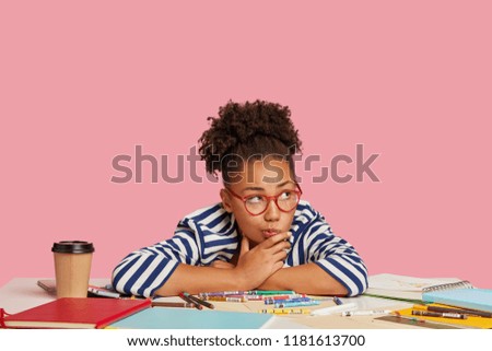 Photo of thoughtful girl multiplier considers about creative ideas, during working process, leans at table with markers, diary, takeaway coffee, look pensively aside, isolated over pink background