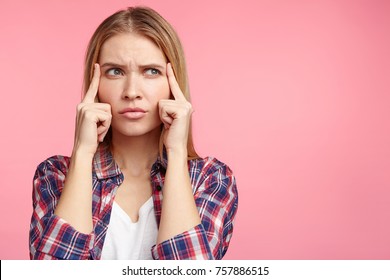 Photo Of Thoughtful Serious Female Model Keeps Fingers On Temples, Tries To Remember Important Information, Can`t Find Quick Answer In Mind, Poses Over Pink Studio With Copy Blank Space For Your Text