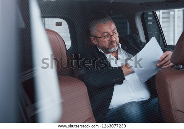 Photo through the glass.\
Paperwork on the back seat of the car. Senior businessman with\
documents.