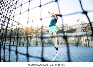 Photo through the net of a paddle tennis court of a female paddle tennis player jumping to hit a ball during a match. Outdoor paddle tennis concept, women playing paddle tennis.