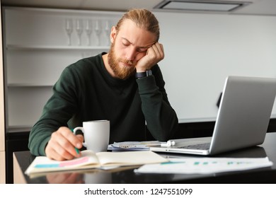 Photo of a tired young bearded man sitting at the kitchen drinking coffee using laptop computer.