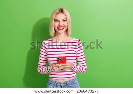 Photo of toothy beaming gorgeous girl with short hairdo dressed striped clothes holding smartphone isolated on green color background