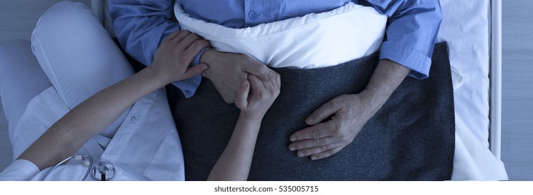 Photo from the top of nurse holding older sick man's hand who lies in hospital bed
