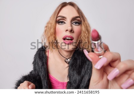 Photo of transgender guy holds lipstick and concentrated at camera has slightly opened mouth dressed in fashionable outfit applies makeup going to have date with boyfriend isolated on white ackground