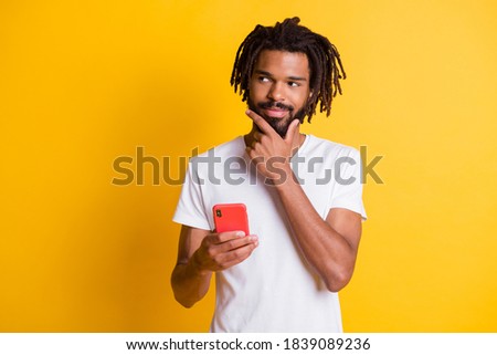Photo of tricky influencer dark skin guy hold telephone look side sly eyes wear t-shirt isolated yellow color background