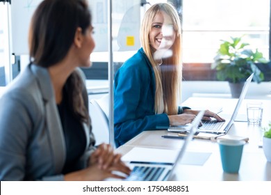 Photo Of Two Business Women Looking At Each Other As They Work With Laptops At The Divided Desk In The Coworking Space. Concept Of Social Distancing.