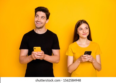 Photo of two fun positive cute nice husband wife people together stylish trendy pondering over what to type holding phones browsing wearing black t-shirt isolated yellow vivid color background