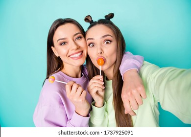Photo of two funny ladies hold lollipop chupa chups hands childish tasty sweets making selfies wear green violet pullovers isolated pastel teal color background