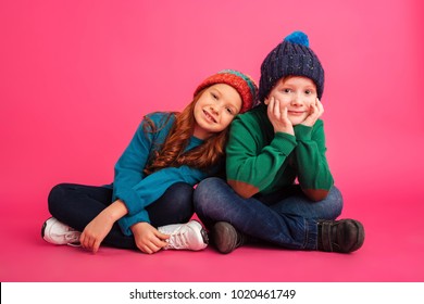 Photo of two happy little children sitting on floor isolated over pink background wearing warm hats. Looking camera.