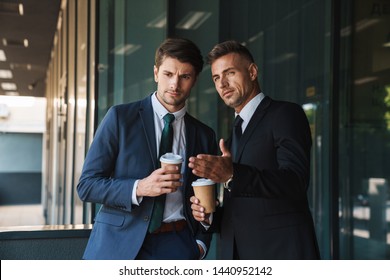 Photo Of A Two Serious Colleagues Handsome Business Men Outdoors At The Street Near Business Center Talking With Each Other Drinking Coffee Gossiping.