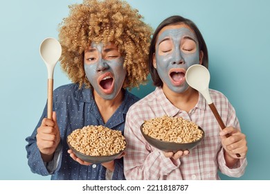 Photo Of Two Women Awake Early In Morning Yawn Going To Eats Cornflakes For Breakfast Applies Beauty Facial Mask Dressed In Slumber Suits Stand Next To Each Other Isolated Over Blue Background