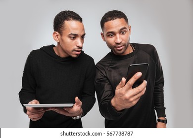 Photo Of Two Young Attractive African Men Standing Over White Background While Using Phone And Tablet Computer.