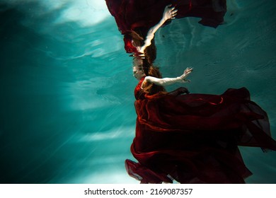 Photo Underwater, A Young Beautiful Woman In Red With Red Hair Reaches For The Surface Of The Water, A Human And His Reflection. Mystical Underwater Portrait