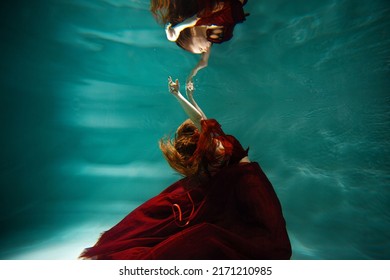 Photo Underwater, A Young Beautiful Woman In Red With Red Hair Reaches For The Surface Of The Water, A Human And His Reflection. Mystical Underwater Portrait