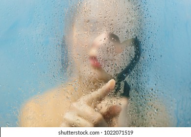 Photo of unrecognizable young woman draws heart on weeping glass shower door, enjoys rest in douche, washes her body, stands behind steam blurred glass with water drops. Rest at bathroom