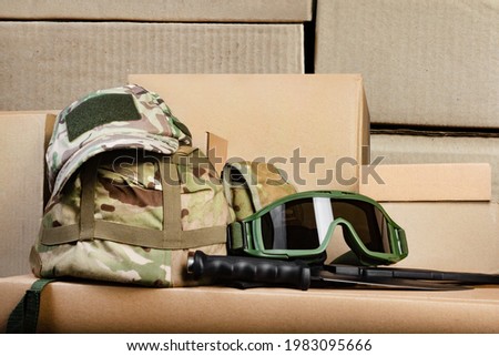 Photo of various military camouflaged armored helmet, cap and soldier googgles.. laying on stacked cardboard boxes.