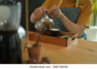 Photo of waiter's hand pouring a hot coffee from a steel kettle to coffee cup on the serving tray full of coffee bean and coffee bean scoop.