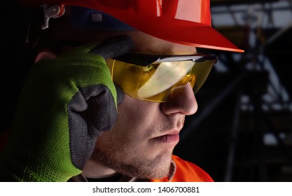 Photo Of A Worker In Orange Jacket Outfit With Protective Gloves And Helmet Putting On Glasses On Construction Area Background.