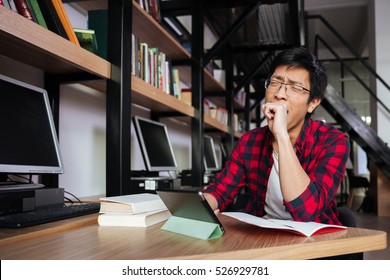 Photo of yawning asian male student dressed in shirt in a cage and wearing glasses using tablet at the library. - Powered by Shutterstock