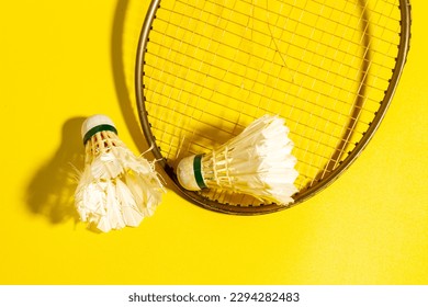 Photo with a yellow rough surface background. Kok or badminton ball or shuttlecock, the condition of the feather is damaged, dirty, with a broken badminton racket and hard shadows on the surface