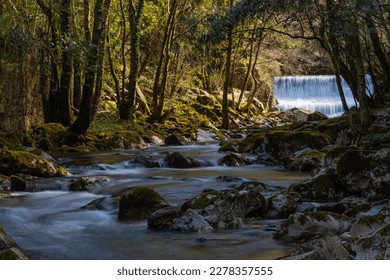 In the photo you can see a silky river running between the rocks and with a curtain waterfall in the background, with the sun's rays entering through the trees. - Shutterstock ID 2278357555