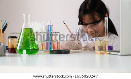Photo of young adorable girl in safety glasses and gloves writing a experiment result while doing a scientist experiment at the modern laboratory with isolated white background.