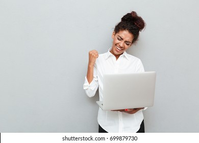 Photo Of Young African Cheerful Business Woman Standing Over Grey Wall With Laptop Computer. Looking Aside Make Winner Gesture.