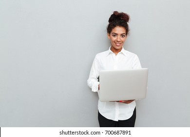 Photo of young african cheerful business woman standing over grey wall with laptop computer. Looking camera.