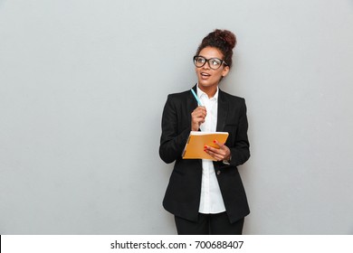 Photo Of Young African Thinking Business Woman Over Grey Wall Looking Aside Writing Notes.