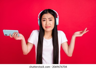 Photo Of Young Asian Girl Unhappy Moody Uncertain Dislike Music Earphones Use Cellphone Isolated Over Red Color Background