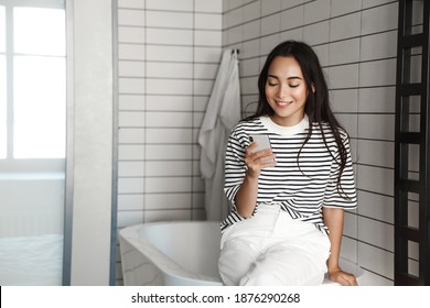 Photo of young asian woman sitting in bathroom with smartphone. Girl using phone and sit on bathtub, smiling and reading message. - Powered by Shutterstock