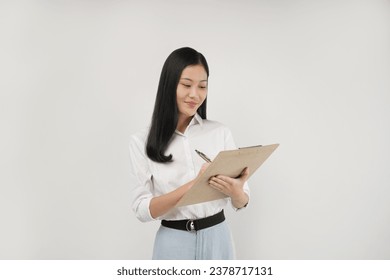 Photo of young asian woman taking notes with pen on clipboard, looking happy, standing against white background