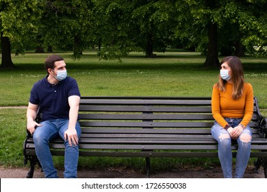 Photo of a young and attractive couple sitting on a bench.  They are wearing a face mask and talking with social distancing in the park - Powered by Shutterstock