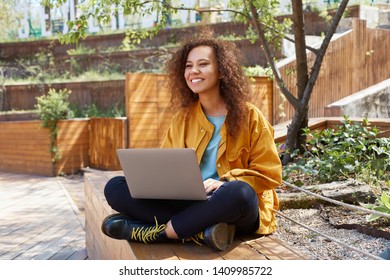 Photo of young attractive dark skinned curly blogger lady, sits in the park with legs crossed, with a laptop on lap, looks away and create a new blog topic.
