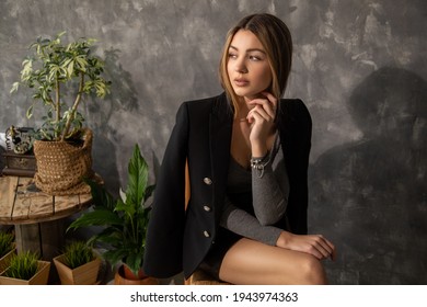 Photo Of Young Beautiful Business Woman Designer Architect Student Sits On Chair Against Background Of Concrete Wall, Wooden Furniture, Green Plant