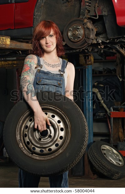 Photo of a young beautiful redhead mechanic\
wearing overalls and holding a wheel.  Attached property release is\
for arm tattoos.