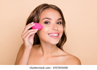 Photo of young beautiful smiling woman applying foundation on cheek with beauty blender isolated on beige color background